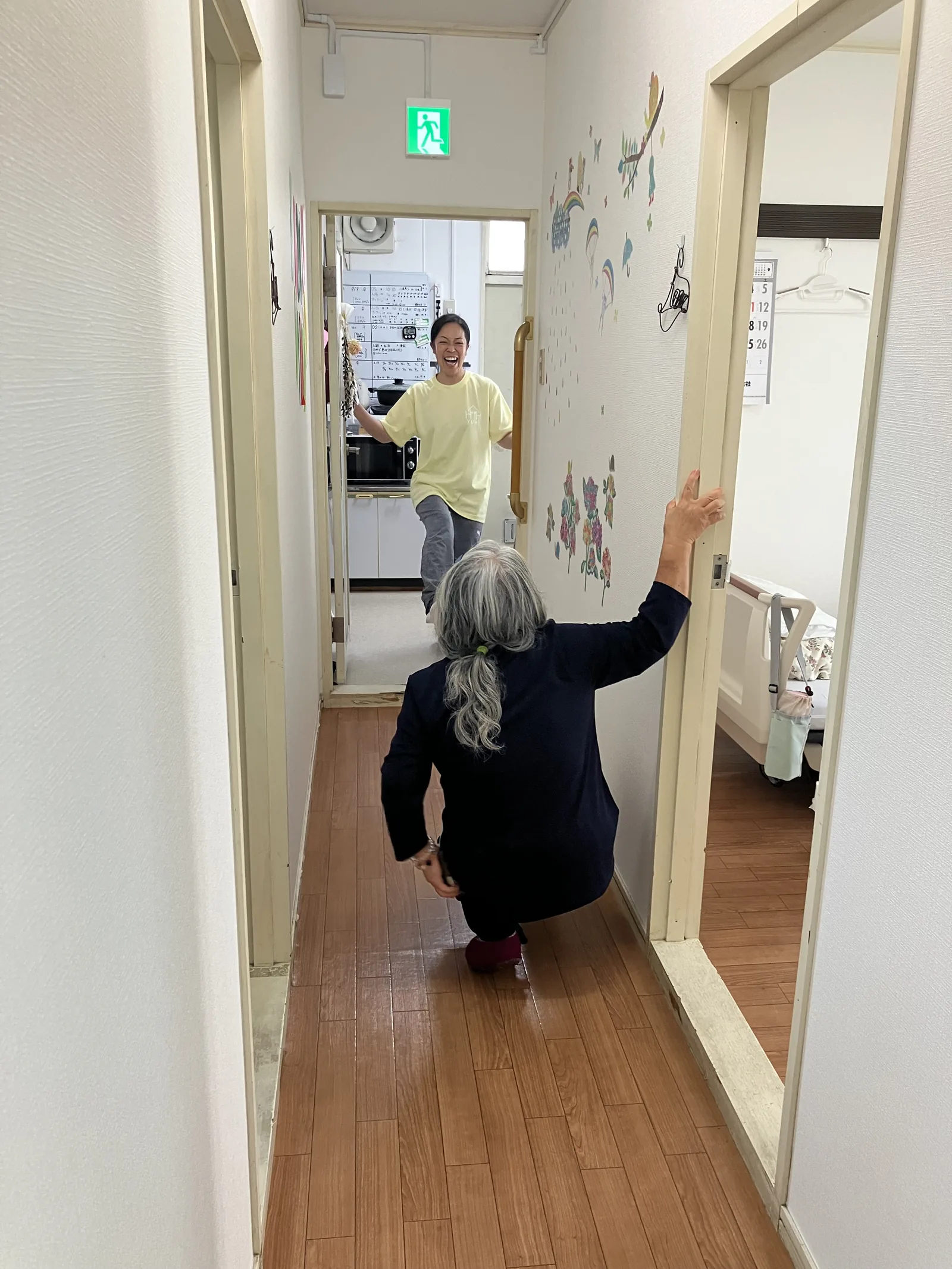 ホームの廊下で歩行練習をする入居者を、スタッフが両手を広げて迎えている様子